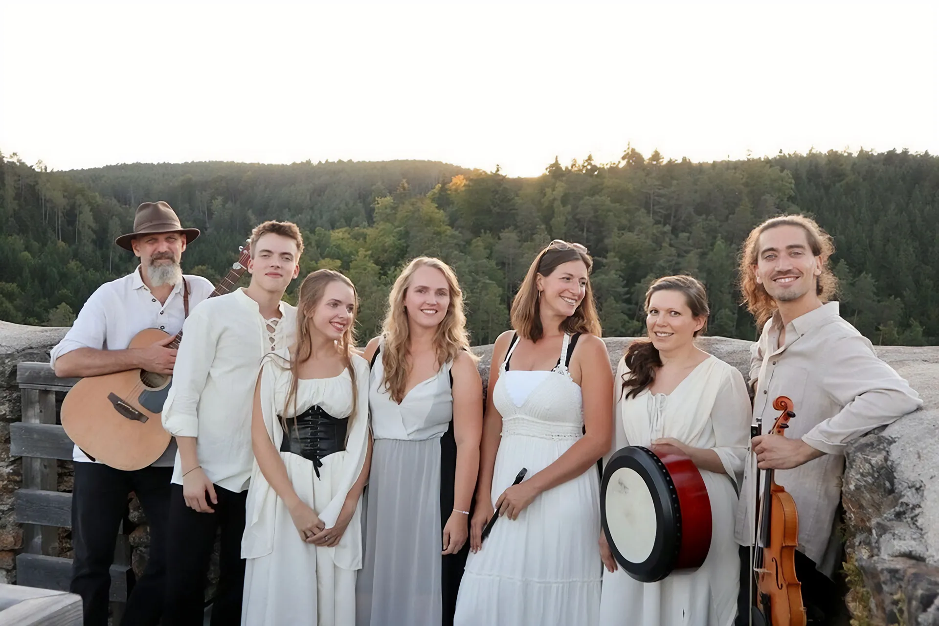 Die siebenköpfige Band und Irish Dance Group in heller Tracht auf einer Aussichtsplattform über bewaldeten Hügeln